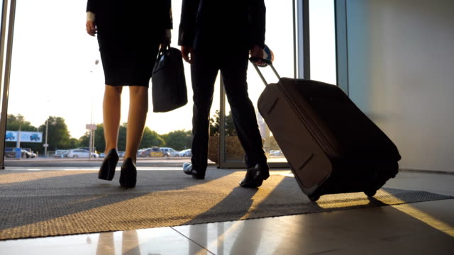 Business-man-and-woman-with-luggage-going-from-the-airport-to-city-street.-Follow-to-young-businessman-carrying-suitcase-on-wheels-and-walking-with-his-female-colleague-from-terminal-hall.-Rear-view-Close-up