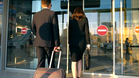 Business-man-and-woman-going-to-the-airport-with-their-luggage.-Young-businessman-carrying-suitcase-on-wheels-and-walking-with-his-female-colleague-to-terminal-hall.-Trip-or-travel-concept.-Rear-view
