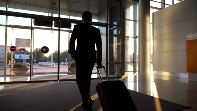 Unrecognizable-man-walking--through-glass-automatic-door-of-modern-airport-to-city-street-and-pulling-suitcase-on-wheels.-Businessman-going-from-terminal-to-cars-parking-with-his-luggage.-Rear-view
