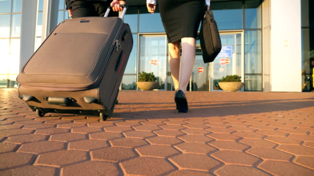 Business-Mann-und-Frau-zum-Flughafen-mit-ihrem-Gepäck.-Junge-Unternehmer-tragen-Koffer-auf-Rädern-und-zu-Fuß-mit-seiner-Kollegin-zu-terminal-Halle.-Oder-einer-Erholungsreise-Konzept.-Ansicht-von-hinten