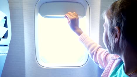 Close-up.-Kind-Mädchen-senkt-das-Bullauge-Vorhang-in-der-Flugzeugkabine,-von-dort-strahlt-ein-helles-Licht.-Mädchen-auf-der-Suche-durch-Flugzeug-Fenster-anzeigen,-Himmel-und-Wolken-und-Landschaft-unter