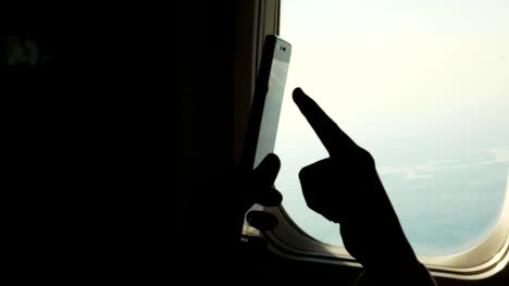 close-up.-dark-silhouette-of-kid-hands-and-mobile-phone-against-airplane's-illuminator.-Child-using,-plays-game-on-smartphone,-tablet-phone-in-airplane-cabin.-passengers-use-gadgets-during-the-flight.-Flight-entertainment-for-kids