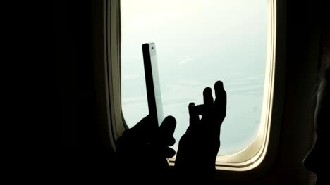 close-up.-dark-silhouette-of-kid-hands-and-mobile-phone-against-airplane's-illuminator.-Child-using,-plays-game-on-smartphone,-tablet-phone-in-airplane-cabin.-passengers-use-gadgets-during-the-flight.-Flight-entertainment-for-kids