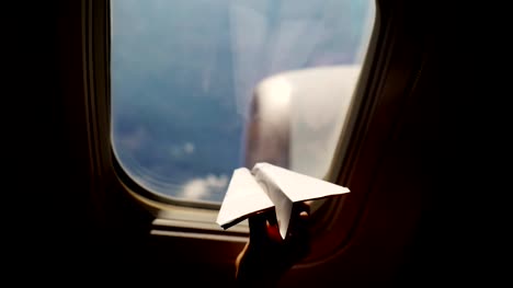 Close-up.-Silhouette-einer-Kinderhand-mit-kleinen-Papierflieger-vor-dem-Hintergrund-der-Flugzeugfenster.-Kind-Flugzeug-Fenster-sitzen-und-spielen-mit-kleinen-Papierflieger.-während-des-Fluges-im-Flugzeug