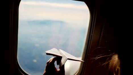 close-up.-Silhouette-of-a-child's-hand-with-small-paper-plane-against-the-background-of-airplane-window.-Child-sitting-by-aircraft-window-and-playing-with-little-paper-plane.-during-flight-on-airplane