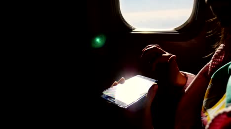 close-up.-the-sun's-rays-are-reflected-in-the-phone.-dark-silhouette-of-kid-hands-and-mobile-phone-against-airplane's-illuminator.-Child-using,-plays-game-on-smartphone,-tablet-phone-in-airplane-cabin