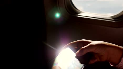 Close-up.-los-rayos-del-sol-se-reflejan-en-el-teléfono.-silueta-oscura-de-las-manos-del-niño-y-el-teléfono-móvil-contra-iluminador-del-avión.-Niño-con,-juego-de-juegos-en-smartphone,-teléfono-tableta-en-cabina-de-avión