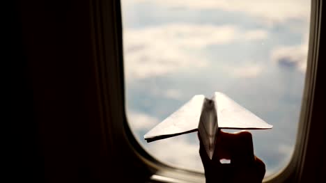 Close-up.-Silhouette-einer-Kinderhand-mit-kleinen-Papierflieger-vor-dem-Hintergrund-der-Flugzeugfenster.-Kind-Flugzeug-Fenster-sitzen-und-spielen-mit-kleinen-Papierflieger.-während-des-Fluges-im-Flugzeug