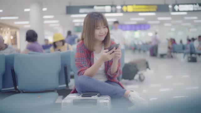 Gerne-Asiatin-mit-und-überprüfen-ihr-Smartphone-sitzend-auf-Stuhl-im-terminal-Halle-während-des-Wartens-ihr-Fluges-am-Abfluggate-im-internationalen-Flughafen.-Frauen-glücklich-im-Flughafenkonzept.