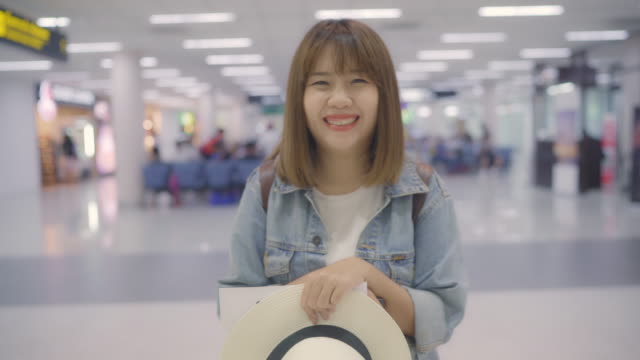 Happy-Asian-woman-smiling-to-camera-while-staying-at-terminal-in-international-airport,-female-tourist-ready-to-travel-on-vacation.-Lifestyle-women-happy-holiday-in-the-airport-concept.