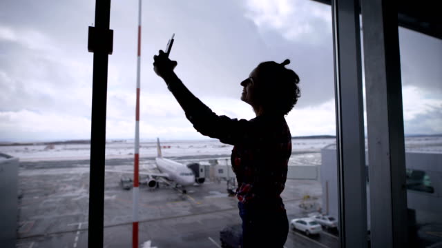 Beautiful-brunette-girl-with-short-hair-at-the-airport.-She-is-happy.-Since-she-was-going-on-vacation-and-plans-to-travel-is-in-a-good-mood.-Standing-panoramic-window-in-the-airport-makes-selfie