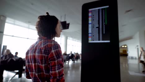 Girl-standing-in-the-airport-and-expects-ads-about-landing-on-the-board-of-the-aircraft.-Brunette-considering-the-scoreboard-with-a-schedule-of-airplanes,-looks-at-his-watch-and-using-her-smartphone