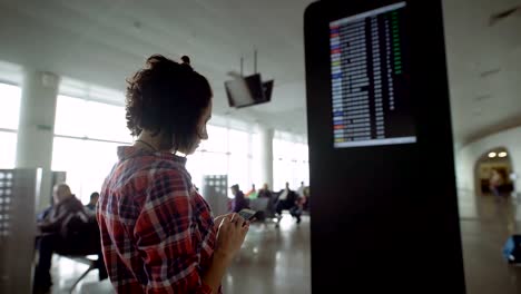 Young-dark-haired-woman-in-chequered-shirt-is-standing-near-information-board-in-the-airport-and-looking-for-information-about-her-departure.-Girl-with-mobile-phone-waiting-her-flight-on-timetable