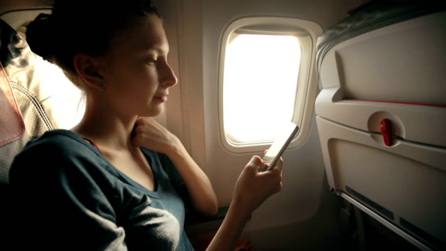Tourist-woman-sitting-near-airplane-window-at-sunset-and-using-mobile-phone-during-flight