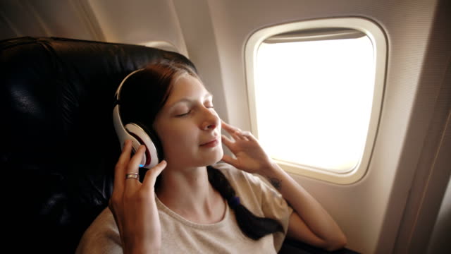 Young-woman-in-wireless-headphones-listening-to-music-and-smiling-during-fly-in-airplane