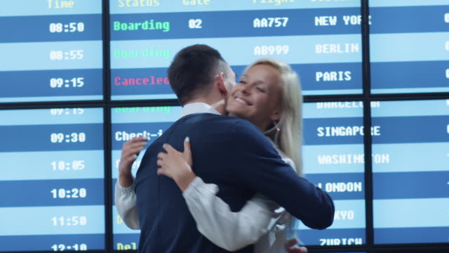 Man-and-Woman-meeting-in-the-Airport-Arrival-Hall-next-to-Information-Board.-People-are-Hugging-each-other-and-Smiling.