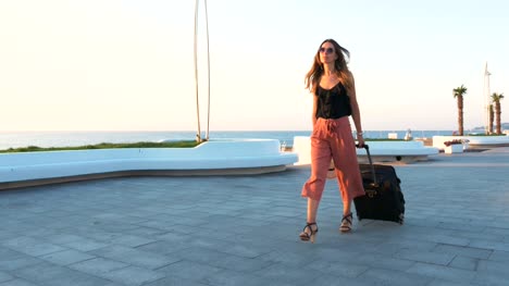 Junge-Geschäftsfrau-zu-Fuß-mit-Koffer-am-Meer-bei-Sonnenuntergang