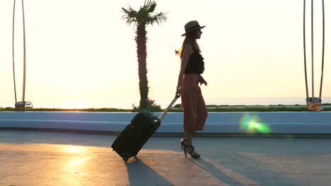 Señora-joven-caminando-con-la-maleta-en-la-playa-durante-el-atardecer