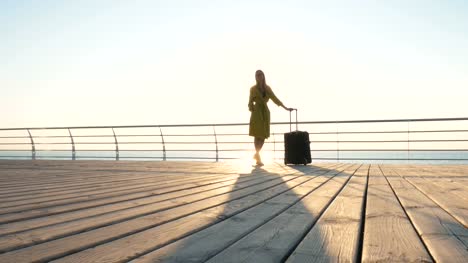 Young-business-lady-with-suitcase-at-the-seafront-during-sunset,-dolly