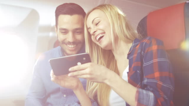 Beautiful-Young-Blonde-with-Handsome-Hispanic-Male-Play-with-Smartphone-on-their-Holiday-Flight.-New-Commercial-Plane-Interior-is-Visible.