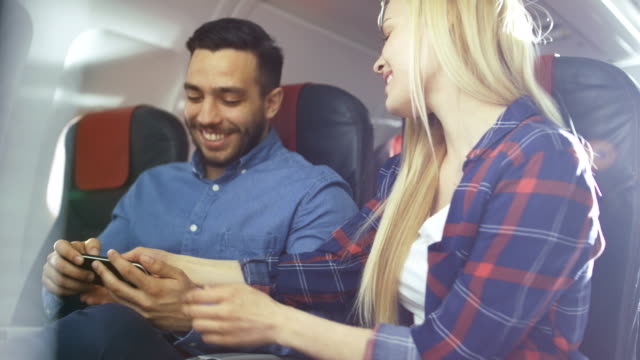 Schöne-junge-Blondine-mit-schöner-Hispanic-männlichen-spielen-mit-Smartphone-auf-ihren-Urlaub-Flug.-Neue-kommerzielle-Flugzeug-Innenraum-ist-sichtbar.
