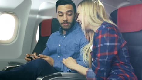 On-a-Board-of-Commercial-Airplane-Beautiful-Young-Blonde-with-Handsome-Hispanic-Male-Watch-Videos-on-Smartphone-and-Smile.-Sun-Shines-Through-Aeroplane-Window.