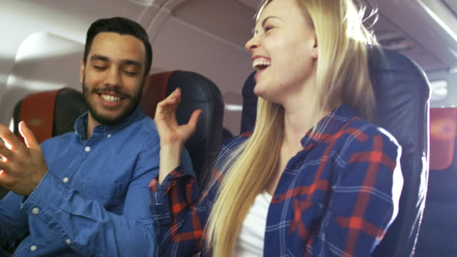 En-un-vuelo-de-avión-comercial-hispano-guapo-narra-divertido-a-su-hermosa-novia-rubia.-Ambos-se-ríen.-Viajan-en-avión-nuevo.