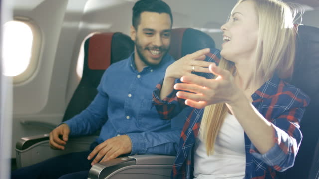 On-a-Commercial-Flight-Beautiful-Blonde-Female-Teaches-Handsome-Hispanic-Man-how-to-Play-Guitar.-Airplane-Is-New,-Sun-Shines-Through-Window.