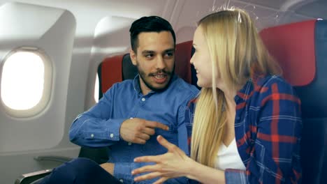 En-un-vuelo-de-avión-comercial-hispano-guapo-narra-divertido-a-su-hermosa-novia-rubia.-Ambos-se-ríen.-Viajan-en-avión-nuevo,-con-el-sol-brillando-a-través-de-la-ventana.