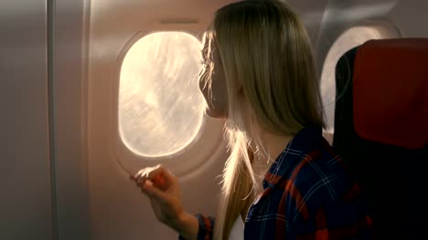 Auf-ein-Flugzeug-schöne-Blonde-Mädchen-öffnet-Schatten-und-schaut-aus-dem-Fenster.-Sonne-ist-Shining-auf-ihr-und-sie-lächelt.