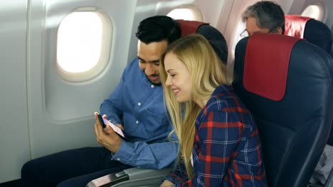 Bei-einem-Board-von-kommerziellen-Flugzeug-schöne-junge-Blondine-mit-schönen-hispanische-männlich-sehen-Social-Media-auf-Smartphone-und-lachen.-Ältere-Passagiere-liest-Buch.