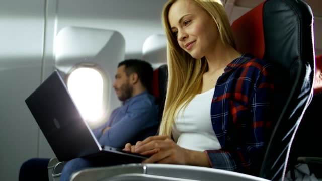 An-Bord-der-kommerziellen-Flugzeug-schöne-arbeitet-junge-Blondine-auf-einem-Laptop-während-ihrer-Hispanic-männlichen-Nachbarn-blickt-aus-dem-Fenster.