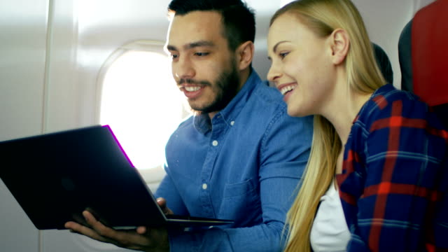 On-a-Board-of-Commercial-Airplane-Beautiful-Young-Blonde-with-Handsome-Hispanic-Male-Watch-Movies-on-a-Laptop-and-Laugh.-Sun-Shines-Through-Aeroplane-Window.