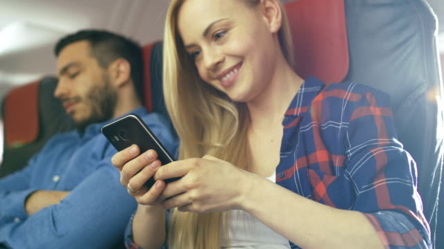 An-Board-des-kommerziellen-Flugzeug-schöne-junge-Blonde-verwendet-Smartphone-während-ihrer-Hispanic-männlichen-Nachbar-schläft.