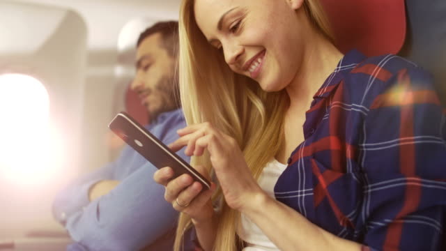 On-Board-of-Commercial-Airplane-Beautiful-Young-Blonde-Uses-Smartphone-while-Her-Hispanic-Male-Neighbor-Sleeps.-Sun-Shines-Through-Plane-Window.