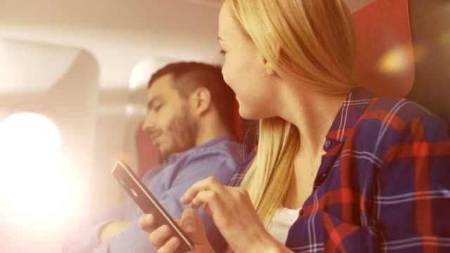 An-Board-des-kommerziellen-Flugzeug-schöne-junge-Blonde-verwendet-Smartphone-während-ihrer-Hispanic-männlichen-Nachbar-schläft.-Sonne-scheint-durch-die-Flugzeugfenster.