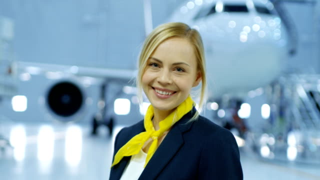 Im-Flugzeug-Wartung-Hangar-junge-schöne-Blonde-Stewardess-/-Flugbegleiter-lächelt-charmant-vor-der-Kamera.-Im-Hintergrund-neue-Flugzeug-ist-sichtbar.