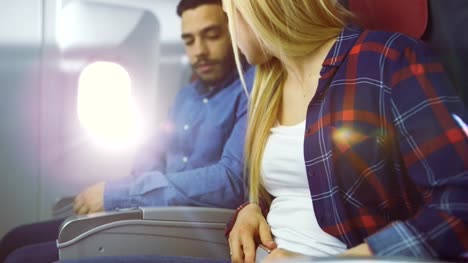 On-a-Plane-Beautiful-Blonde-Female-and-Handsome-Hispanic-Male-Fasten-They're-Seat-Belts-and-Ready-to-Take-off/-Landing.-Sun-Shines-Through-Airplane's-Window.