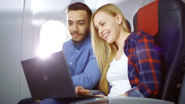 On-a-Board-of-Commercial-Airplane-Beautiful-Young-Blonde-with-Handsome-Hispanic-Male-Watch-Movies-on-a-Laptop-and-Laugh.-Sun-Shines-Through-Aeroplane-Window.