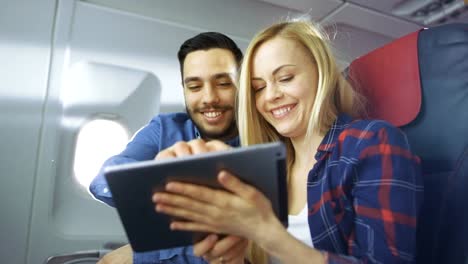 En-un-tablero-de-avión-comercial-hermosa-joven-rubia-con-hermoso-uso-masculino-hispano-Tablet-computadora-y-sonrisa.-El-sol-brilla-a-través-de-la-ventana-del-avión.