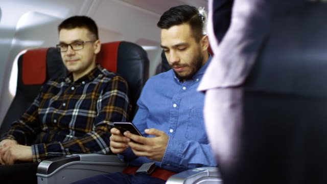 On-a-Commercial-Flight-Young-Hispanic-Male-Uses-Smartphone,-while-His-Neighbor-Looks-out-of-the-Window.-Senior-Man-in-the-Back-Sleeps-Peacefully.