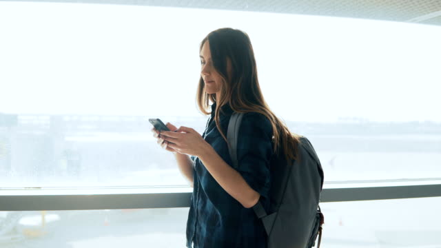 Young-girl-using-smartphone-near-airport-window.-Happy-European-woman-with-backpack-uses-mobile-app-in-terminal.-4K