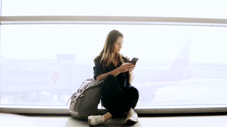Glückliche-Frau-sitzt-mit-Smartphone-Flughafen-Fenster.-Kaukasische-Mädchen-mit-Rucksack-mit-Messenger-app-im-Terminal.-4K