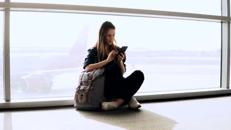 Pretty-girl-sits-using-phone-by-terminal-window.-European-female-passenger-with-backpack-typing-and-smiling-happy.-4K