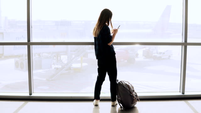 Girl-takes-selfie-near-airport-window,-walks-away.-Happy-Caucasian-female-tourist-using-smartphone-in-terminal-hall.-4K