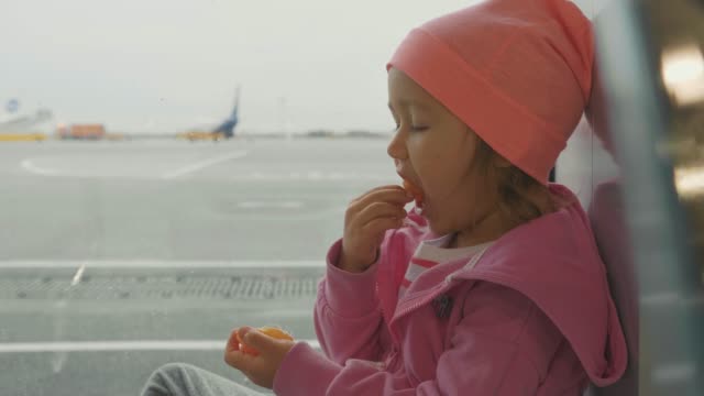 Little-seriously-baby-girl-eats-tangerine-next-to-window-at-airport.