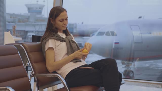 Young-woman-eats-tangerine-at-airport-with-airplane-on-the-background