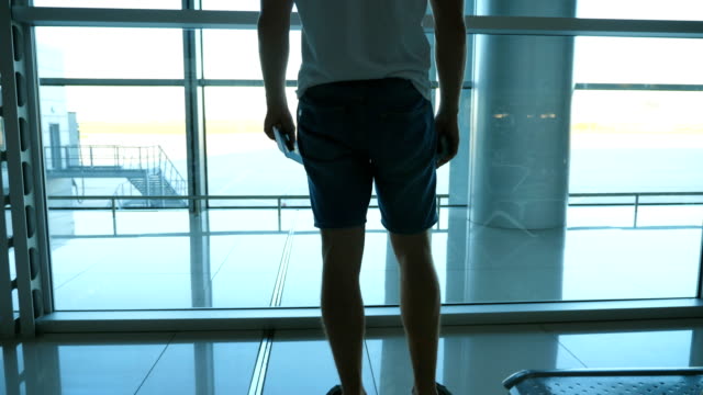 Young-man-with-passport-and-ticket-standing-in-terminal-of-airport-and-looking-at-window.-Handsome-guy-waiting-for-his-airplane.-Rear-back-view-Close-up