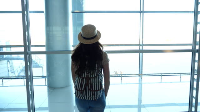 Young-woman-in-hat-with-backpack-coming-to-window-in-terminal-of-airport-and-looking-at-runway.-Girl-waiting-for-her-airplane.-Tourism-and-travel-concept.-Rear-back-view-Close-up