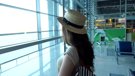 Young-woman-in-hat-with-backpack-standing-in-terminal-of-airport-and-looking-at-window.-Brown-haired-girl-waiting-for-her-airplane.-Tourism-and-travel-concept.-Close-up-Side-view